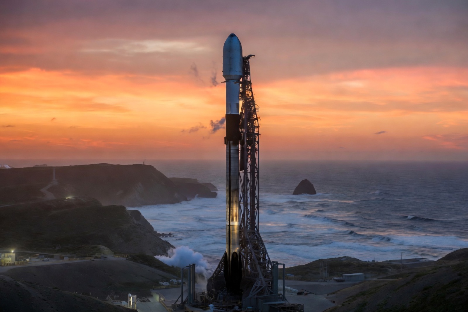 A Falcon 9 rocket vertical on the launch pad at Vandenberg with the Pacific coastline and bluffs in the background under a sunset sky