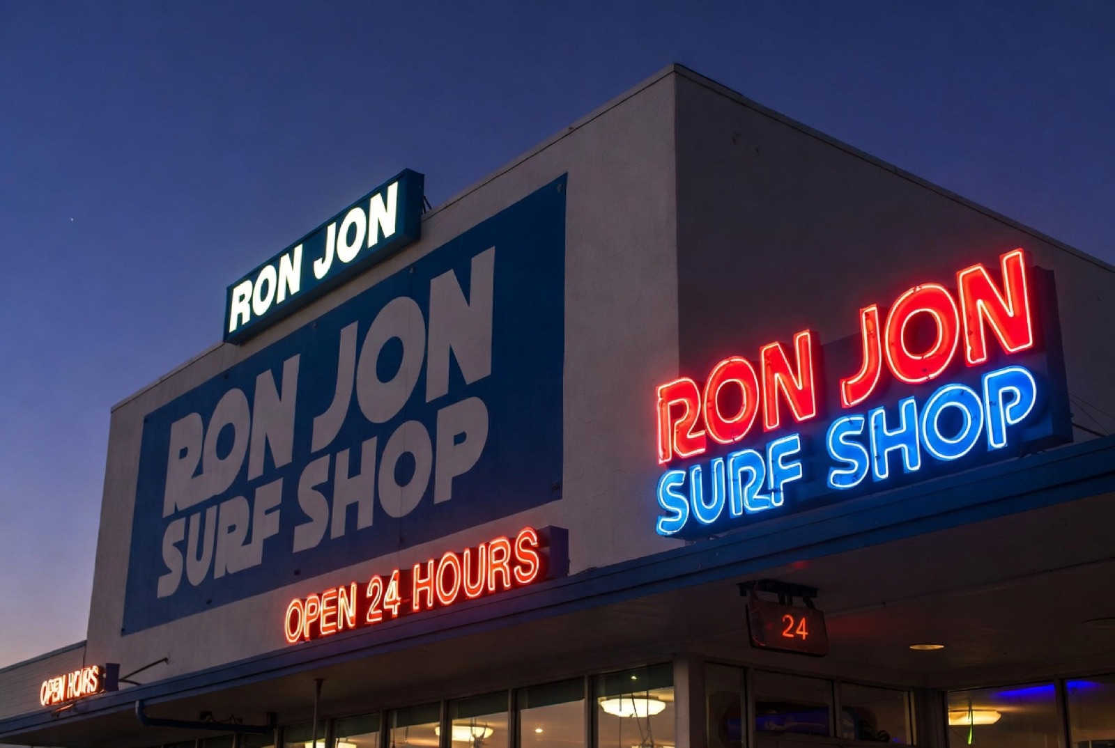 The Ron Jon Surf Shop neon facade at dusk with the iconic 24-hour sign lit up in red and blue